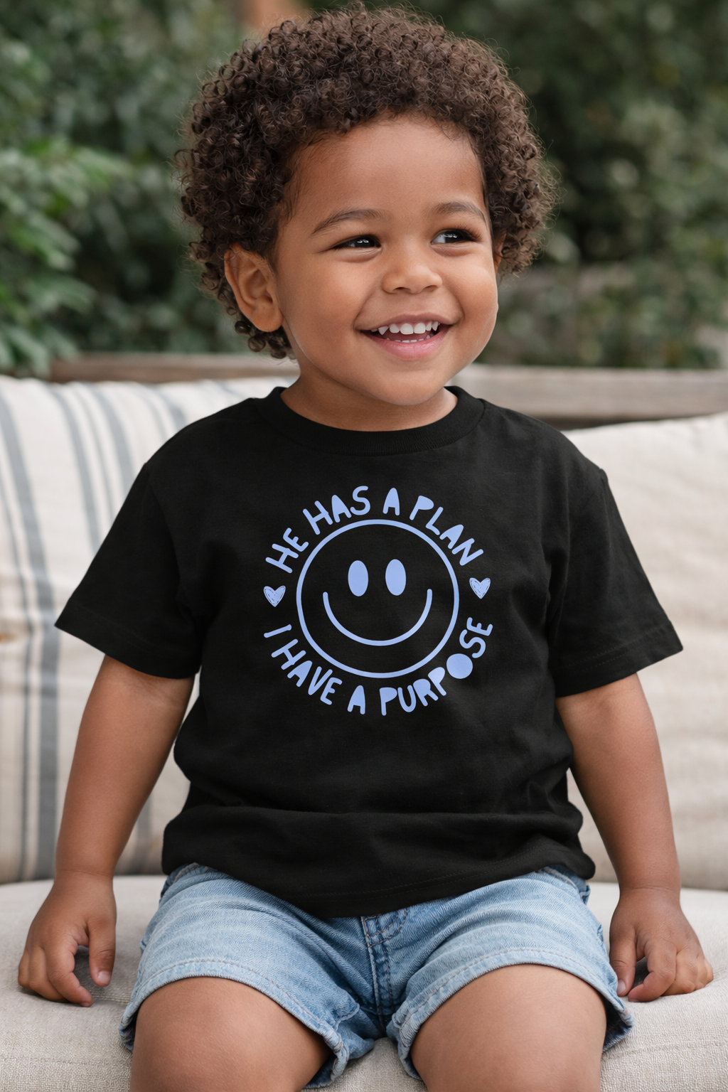 Child wearing a black t-shirt, "he has a plan, I have a purpose," Graphic t-shirt with a smiley face and text, sitting on a couch.